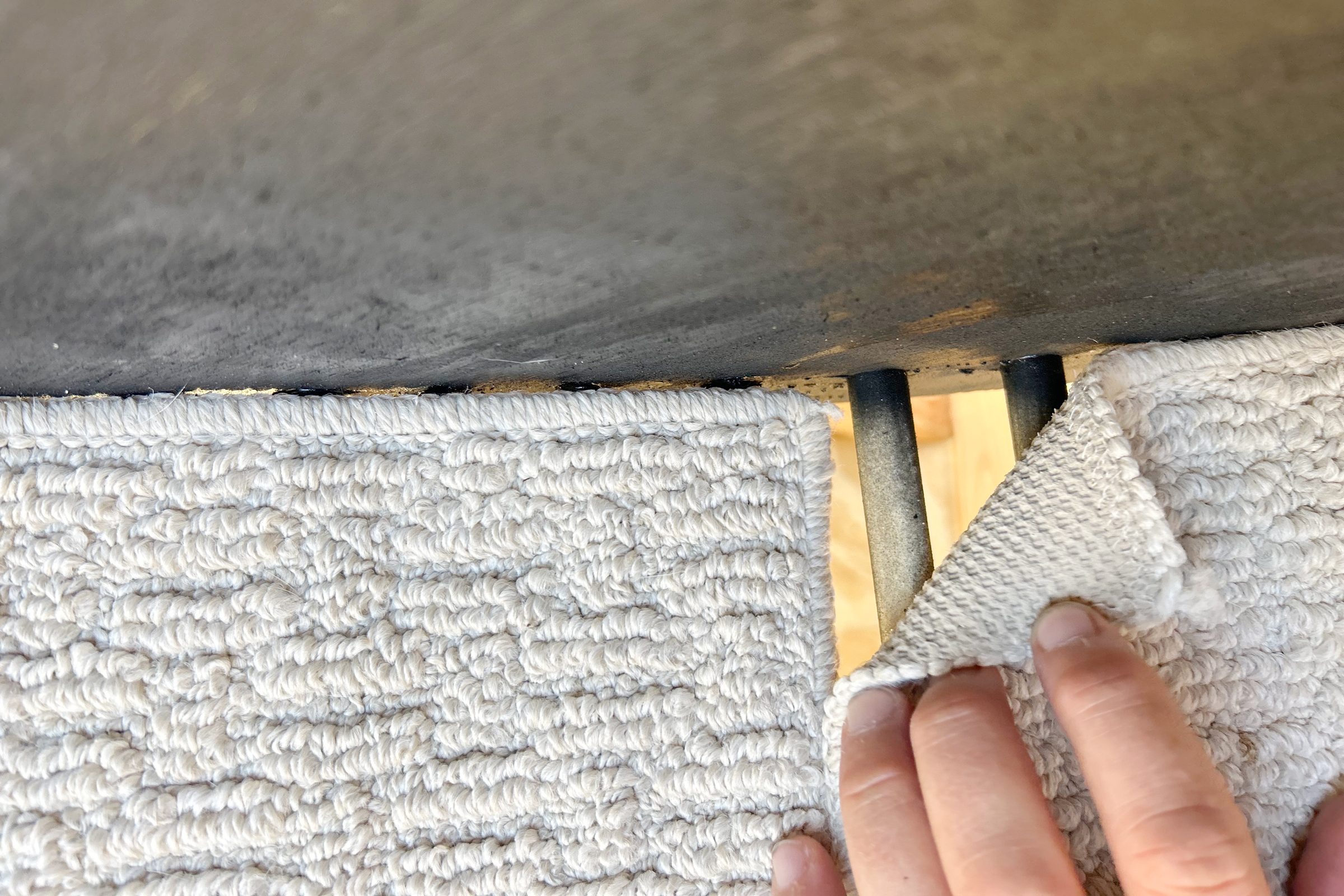 A hand pulls back a section of textured, light-colored fabric, revealing metal bars beneath it, against a wooden surface in a well-lit space.