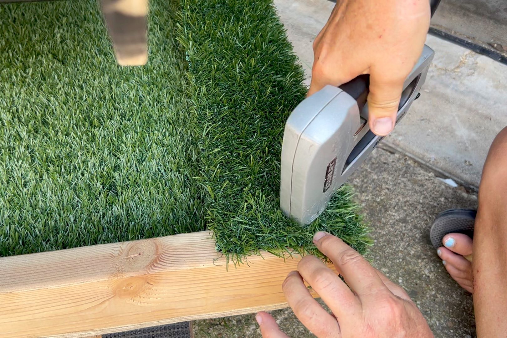 A hand uses a stapler to secure artificial grass onto a wooden frame, positioned on a flat, outdoor surface.