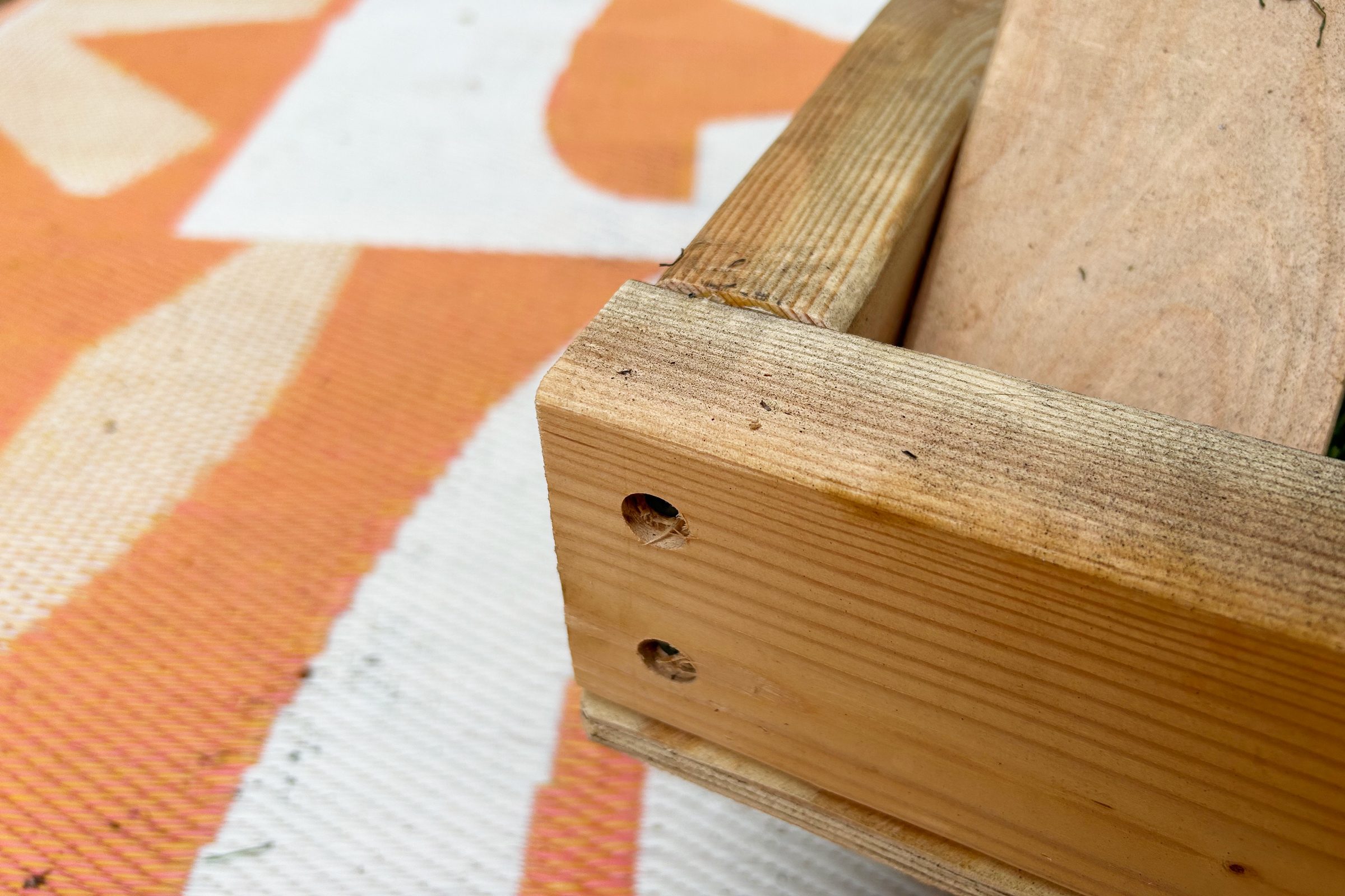 A wooden box with visible nails rests on an orange-patterned surface, highlighting its grain and texture. The background features a simple, light pattern.