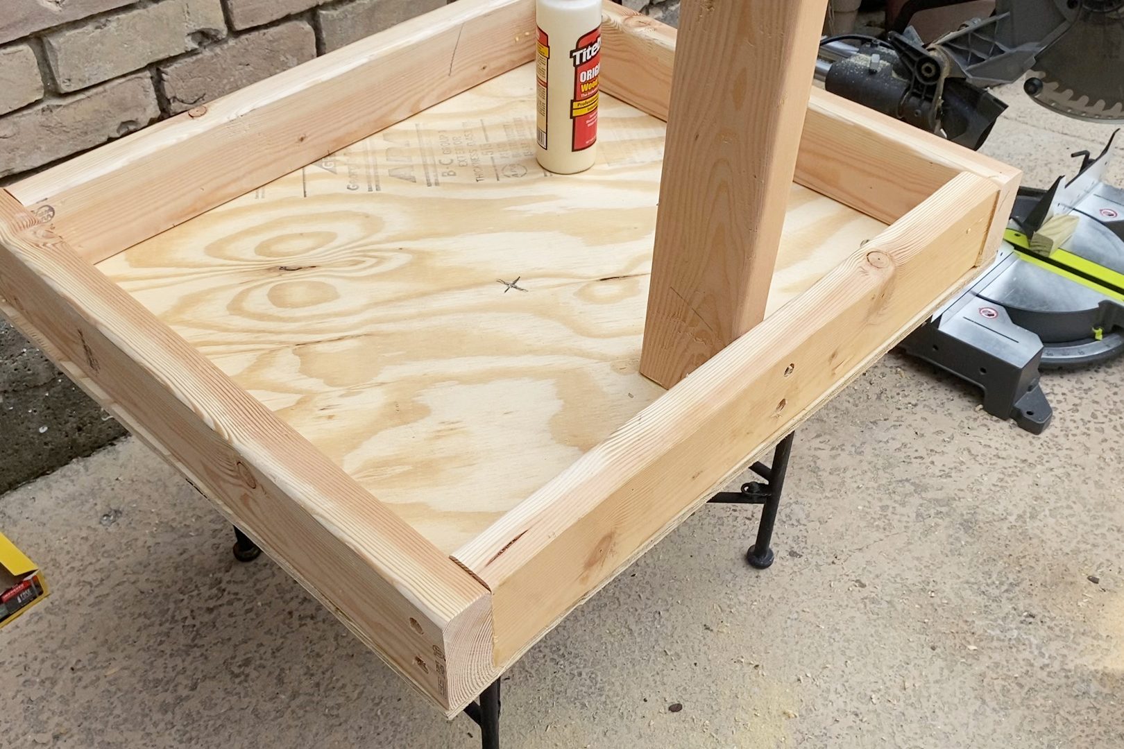 A wooden frame with an attached vertical post is positioned on a work surface, while a bottle of wood glue sits nearby in a outdoor workspace.