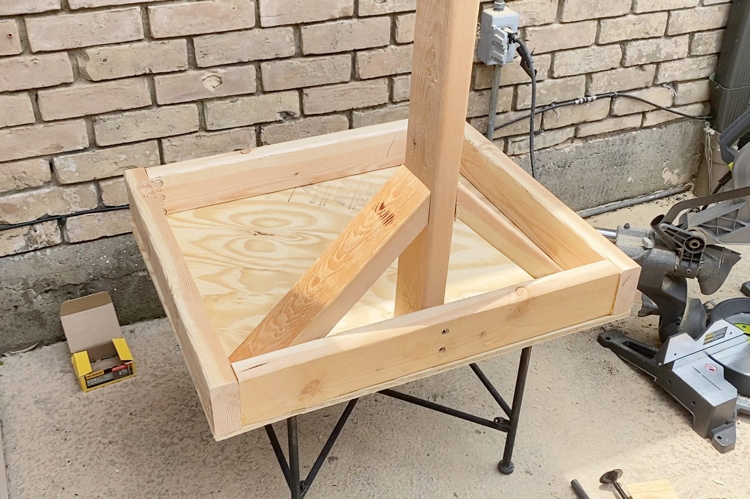 A wooden frame with supports rests on a metal stand in a construction space, surrounded by a brick wall and tools.