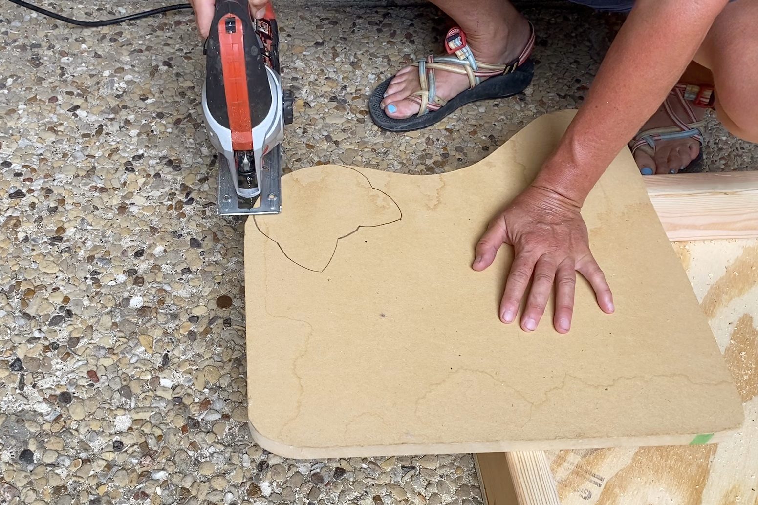 A handsaw is cutting a curved design into a flat piece of wood, resting on a textured outdoor surface, while a hand holds it steady.