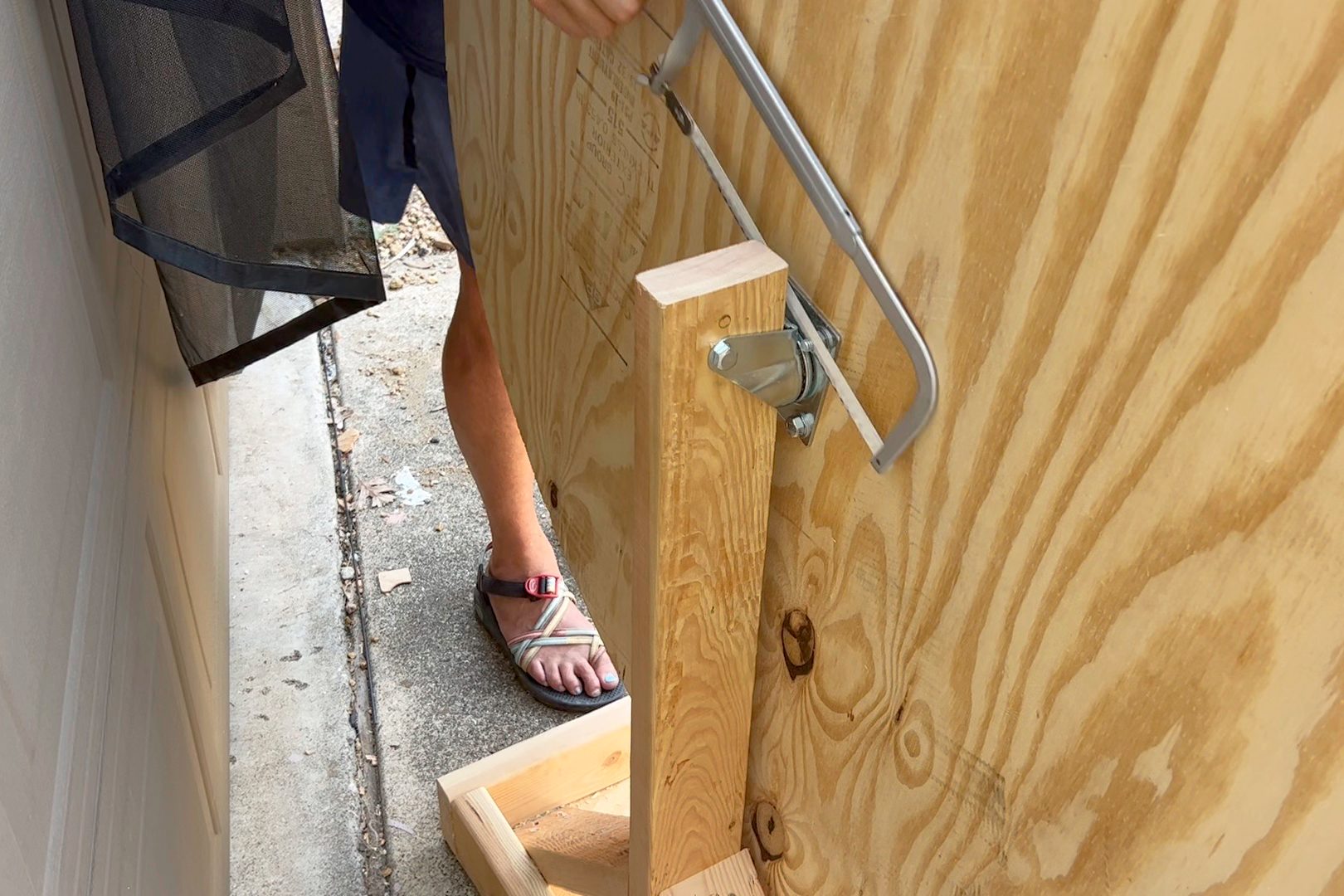 A person is securing a wooden panel using a metallic lever mechanism on a concrete surface, with debris and a nearby wall in the background.