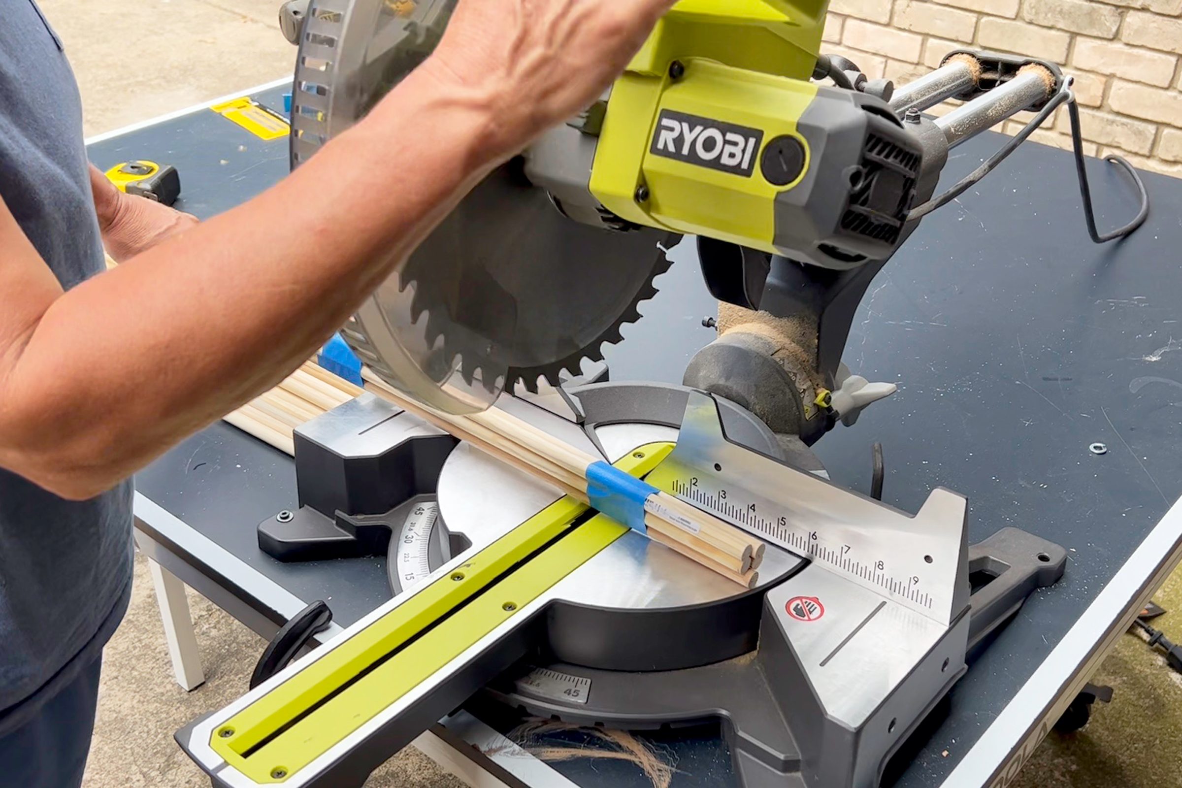 A hand operates a miter saw, cutting wooden pieces secured on a worktable in an outdoor setting, surrounded by various tools and a brick wall.
