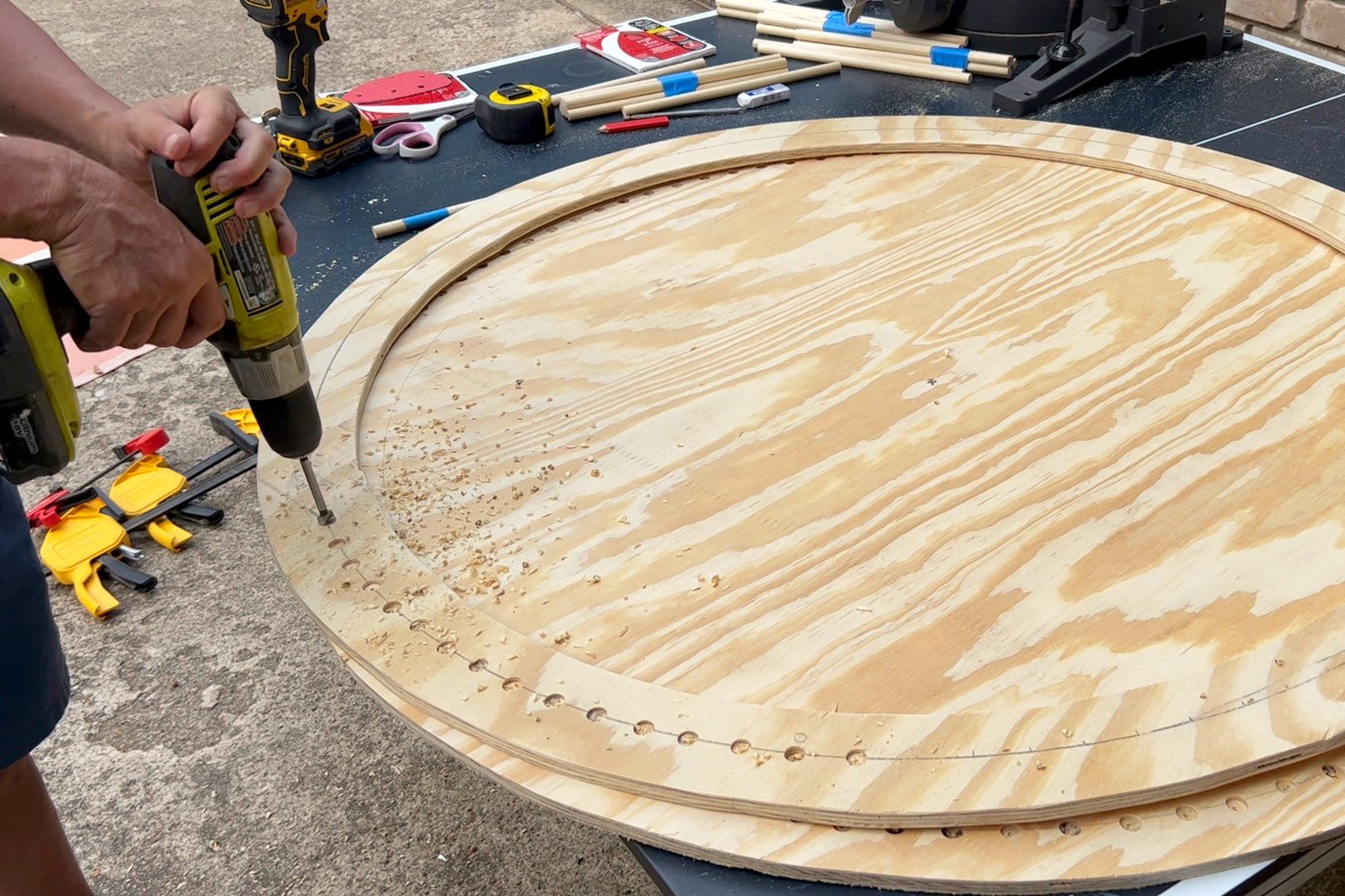 A person drills into a large, circular piece of plywood on a work table, surrounded by various tools and wood scraps on a concrete surface.