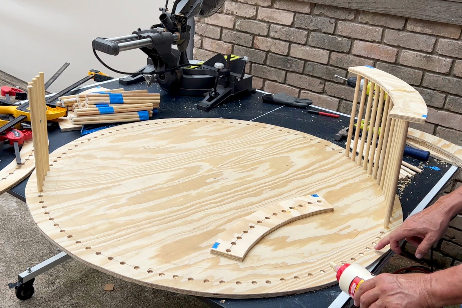 A person is applying glue to wooden pieces on a circular plywood surface, surrounded by tools and materials on a work table near a brick wall.
