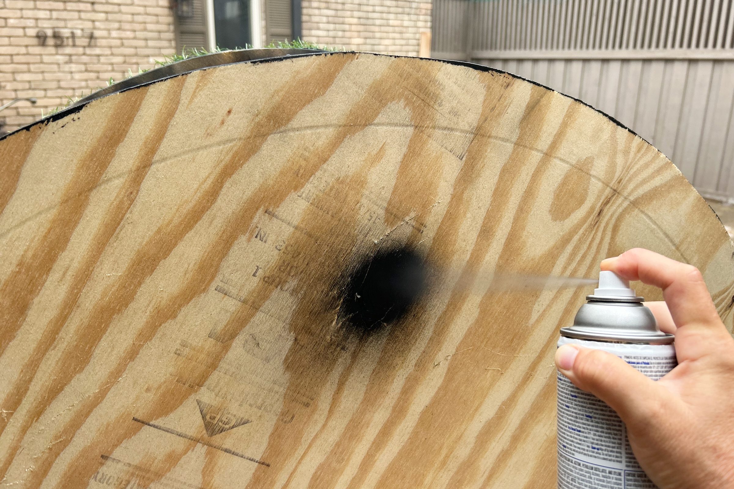 A hand sprays paint onto a wooden circular surface, creating a black spot, with a brick wall and fence visible in the background.