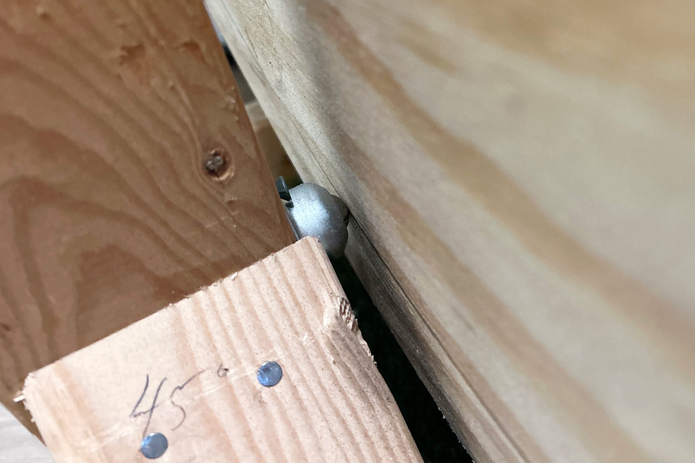 A metal ball joint is positioned between two wooden boards, partially concealed. The surface shows wood grain, with visible markings and nails on one board.