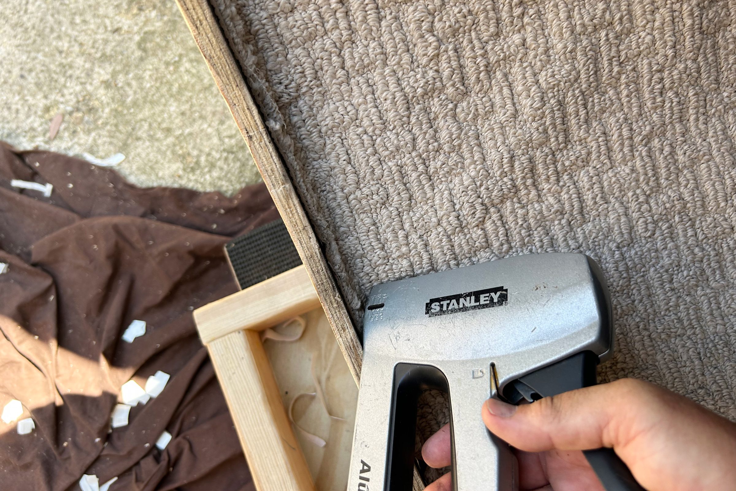 A hand holds a stapler, securing fabric to a wooden frame on a brown cloth, surrounded by pieces of material and a textured carpet.