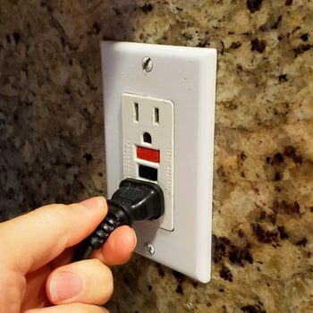 A hand is inserting a black power cord into a white electrical outlet on a textured, light-colored stone wall.