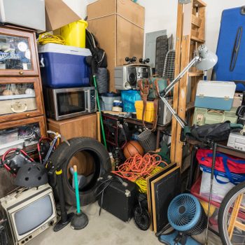 A cluttered garage contains various items like a microwave, bicycle tire, old television, and tools, stacked against walls filled with boxes and bags.
