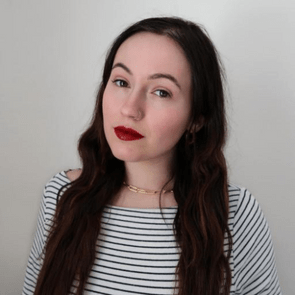 A woman with long brown hair and red lipstick poses calmly, wearing a striped shirt against a neutral wall backdrop.