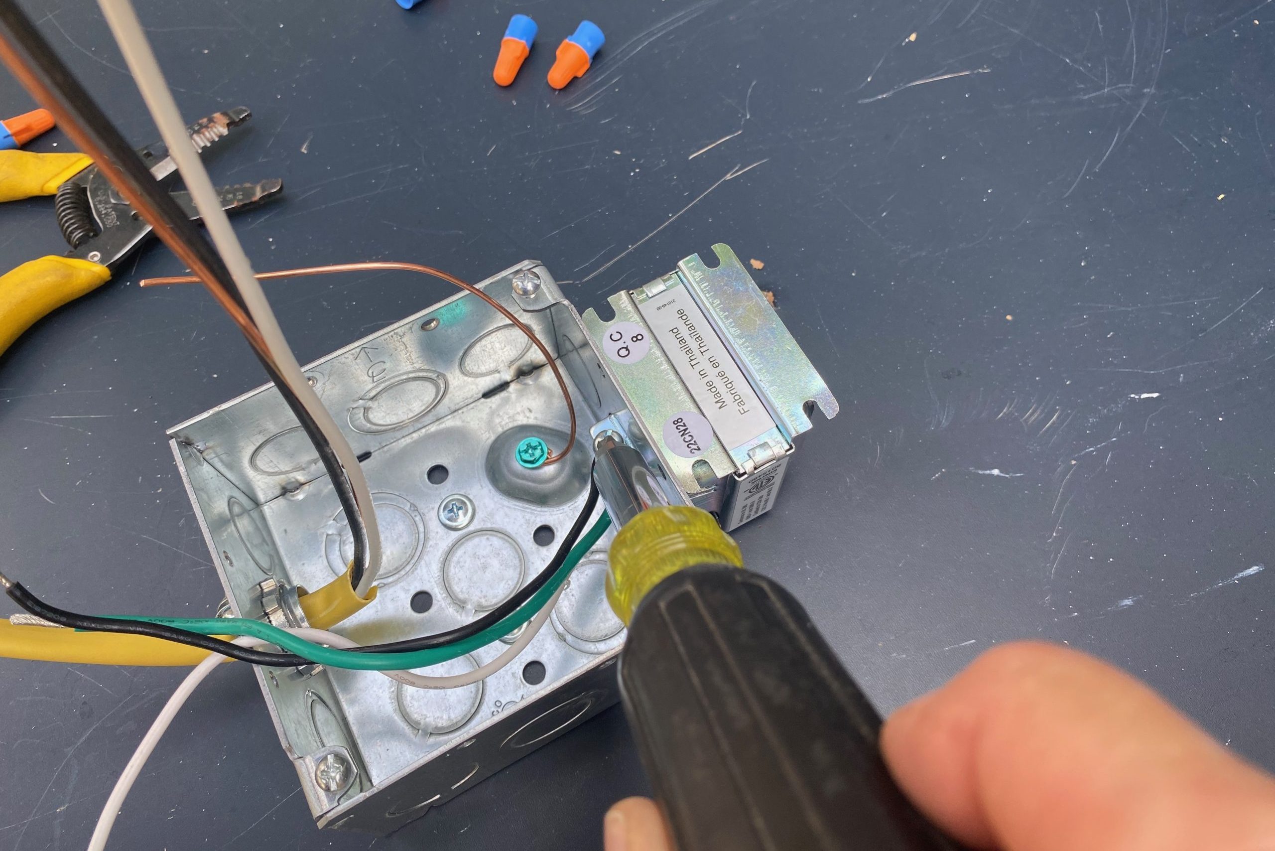 A hand uses a screwdriver to secure wiring connections in a metal electrical box, surrounded by tools and wire connectors on a black surface.