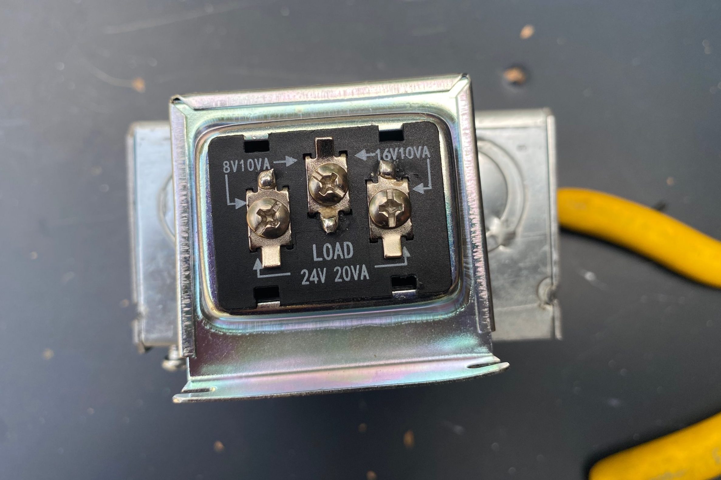 A metal electrical component with three terminal connections displays voltage specifications, placed on a dark surface next to a yellow wire.