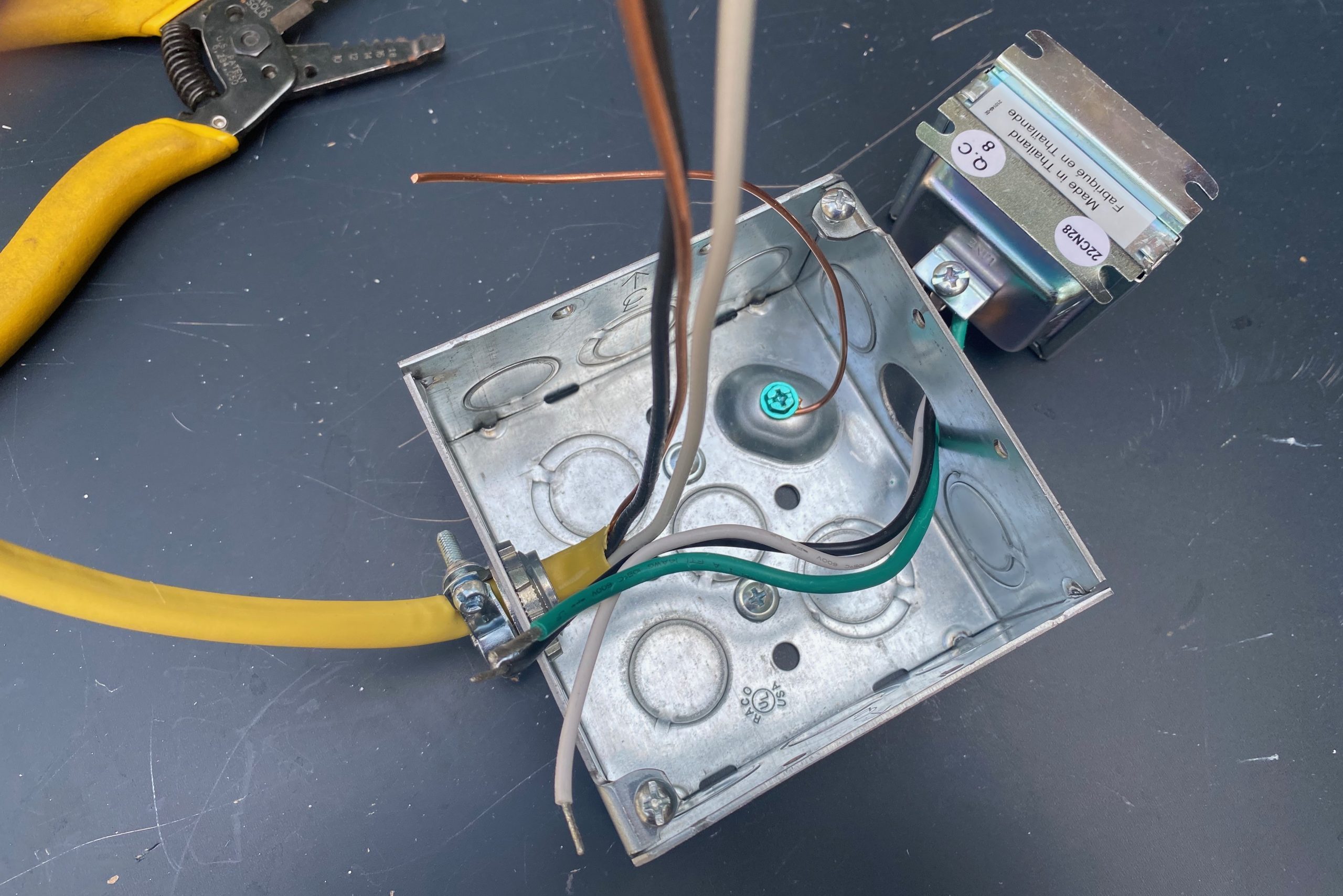 A metal electrical box contains multiple wires, with yellow conduit visible. Nearby, a pair of wire strippers rests on a dark surface.
