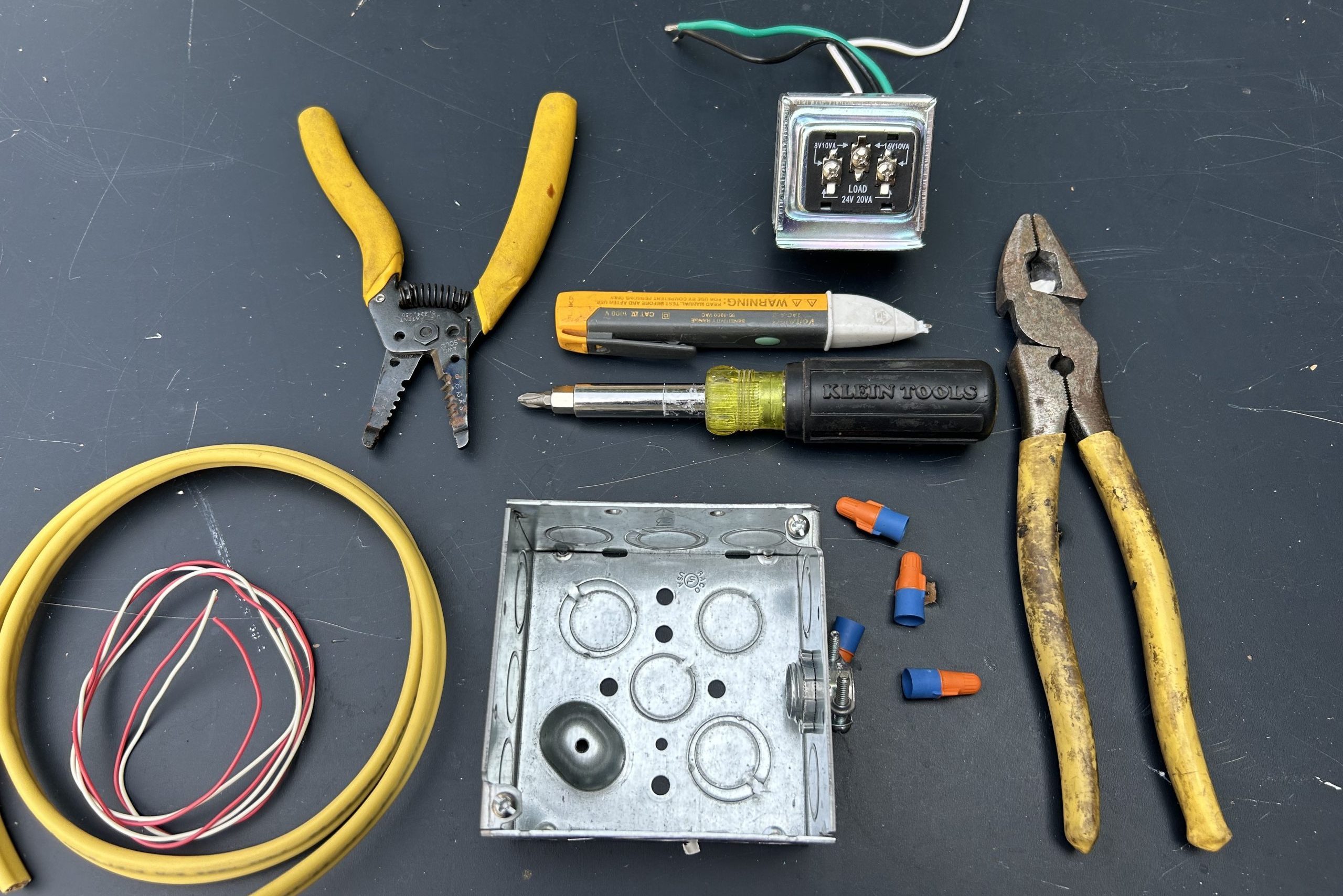 A collection of electrical tools and components is arranged on a dark surface, including wire strippers, pliers, a connector box, and various wires.