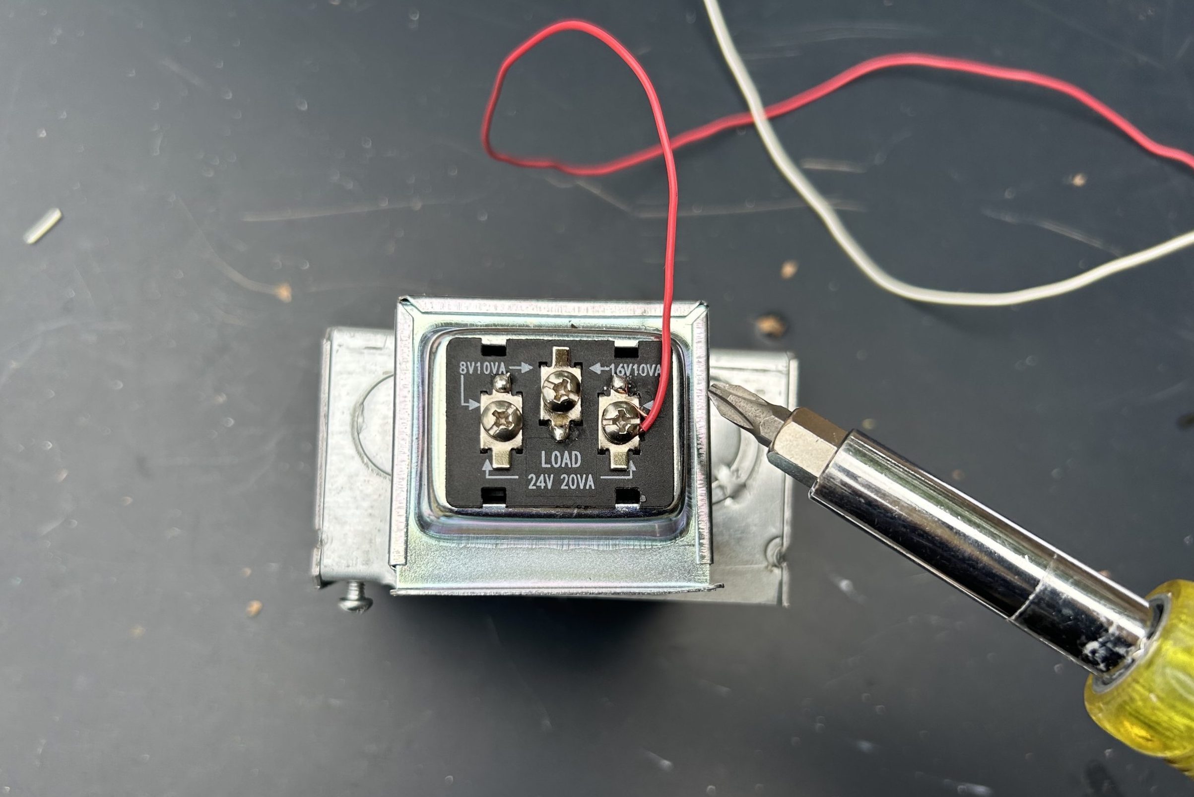 A screwdriver is positioned near a relay with a red wire attached, sitting on a dark surface scattered with small debris.