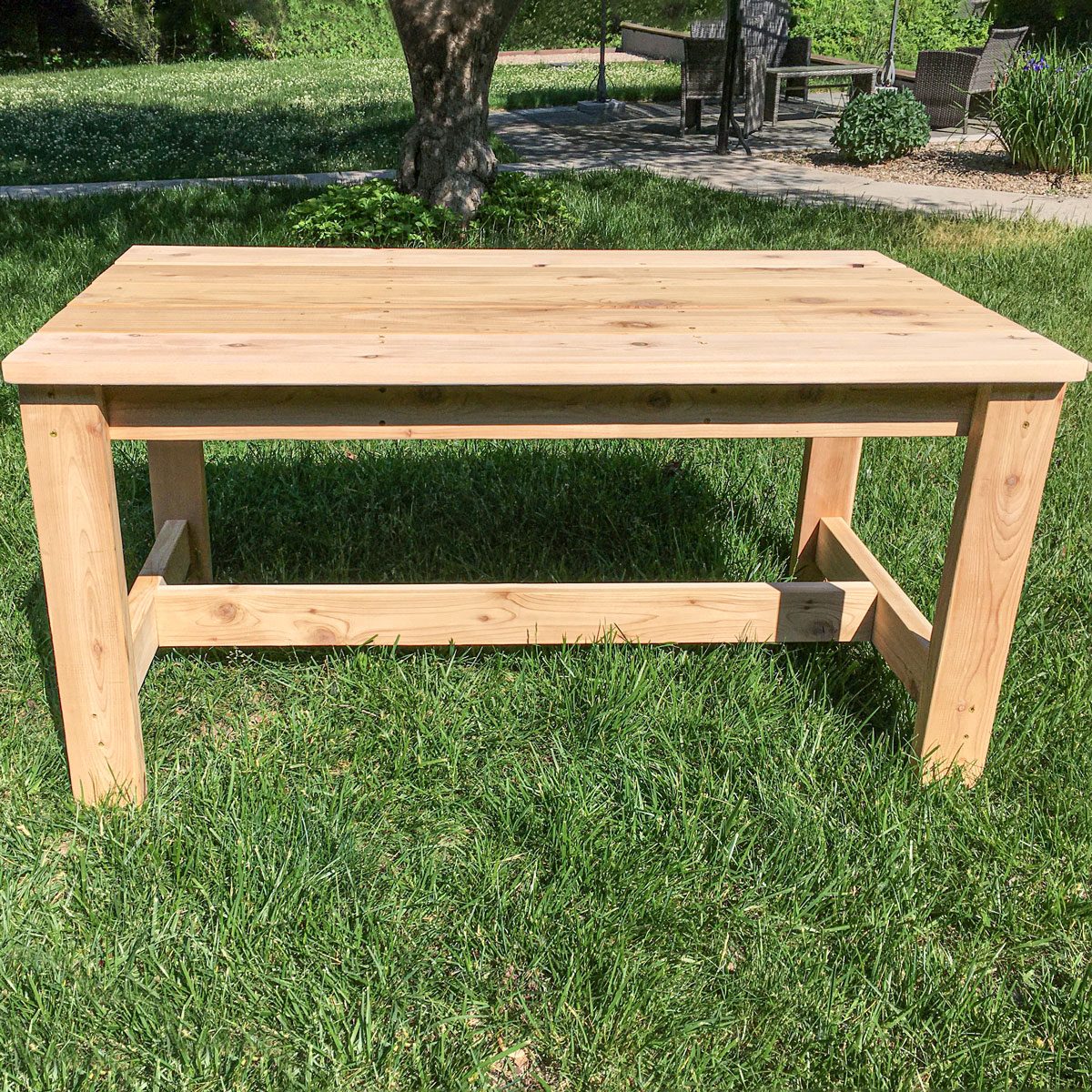 A wooden table stands on grass, showcasing its smooth surface and sturdy legs, amidst a garden setting with trees and distant furniture.