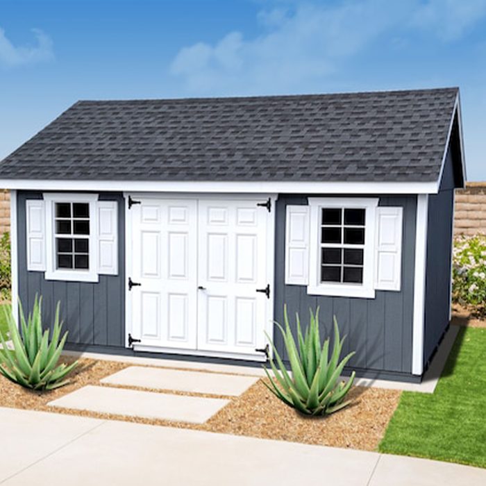 A small, dark-gray shed with white trim features double doors and windows, surrounded by gravel and greenery on a sunny day.