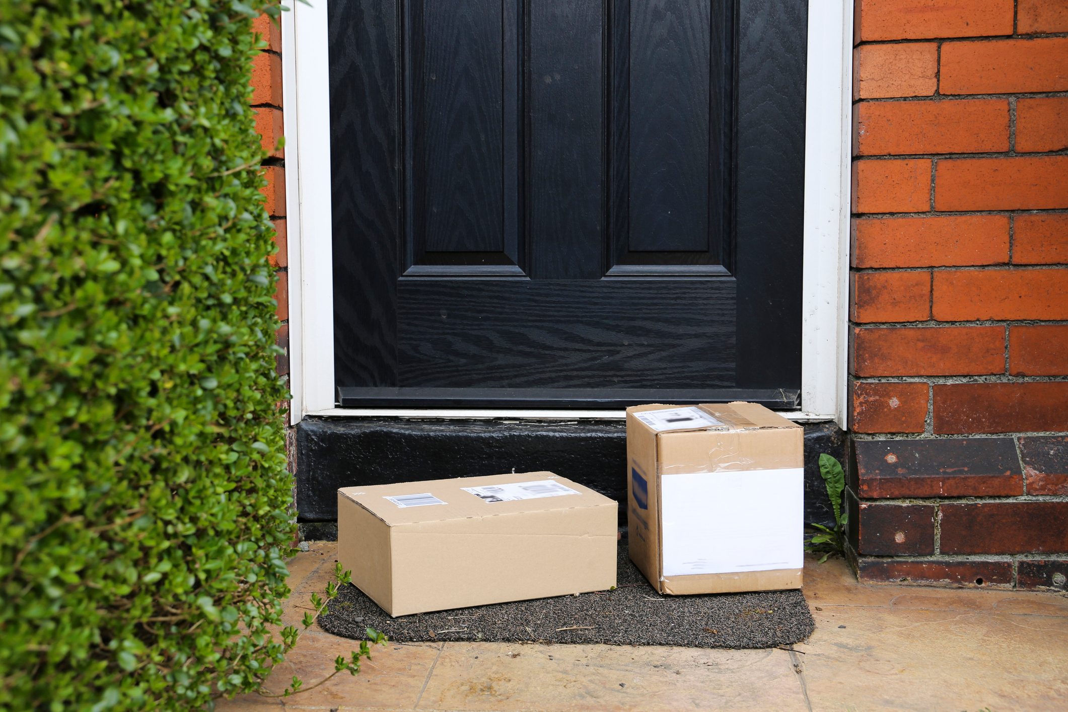 Two packages rest on a doormat in front of a black door, surrounded by a bush and brick wall, suggesting recent delivery.