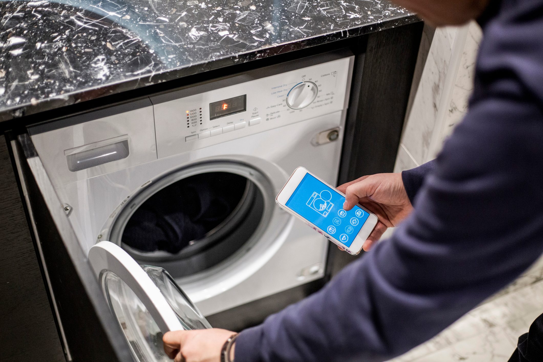A person holds a smartphone while opening a washing machine door to check the laundry, set against a marble countertop and dark cabinetry.