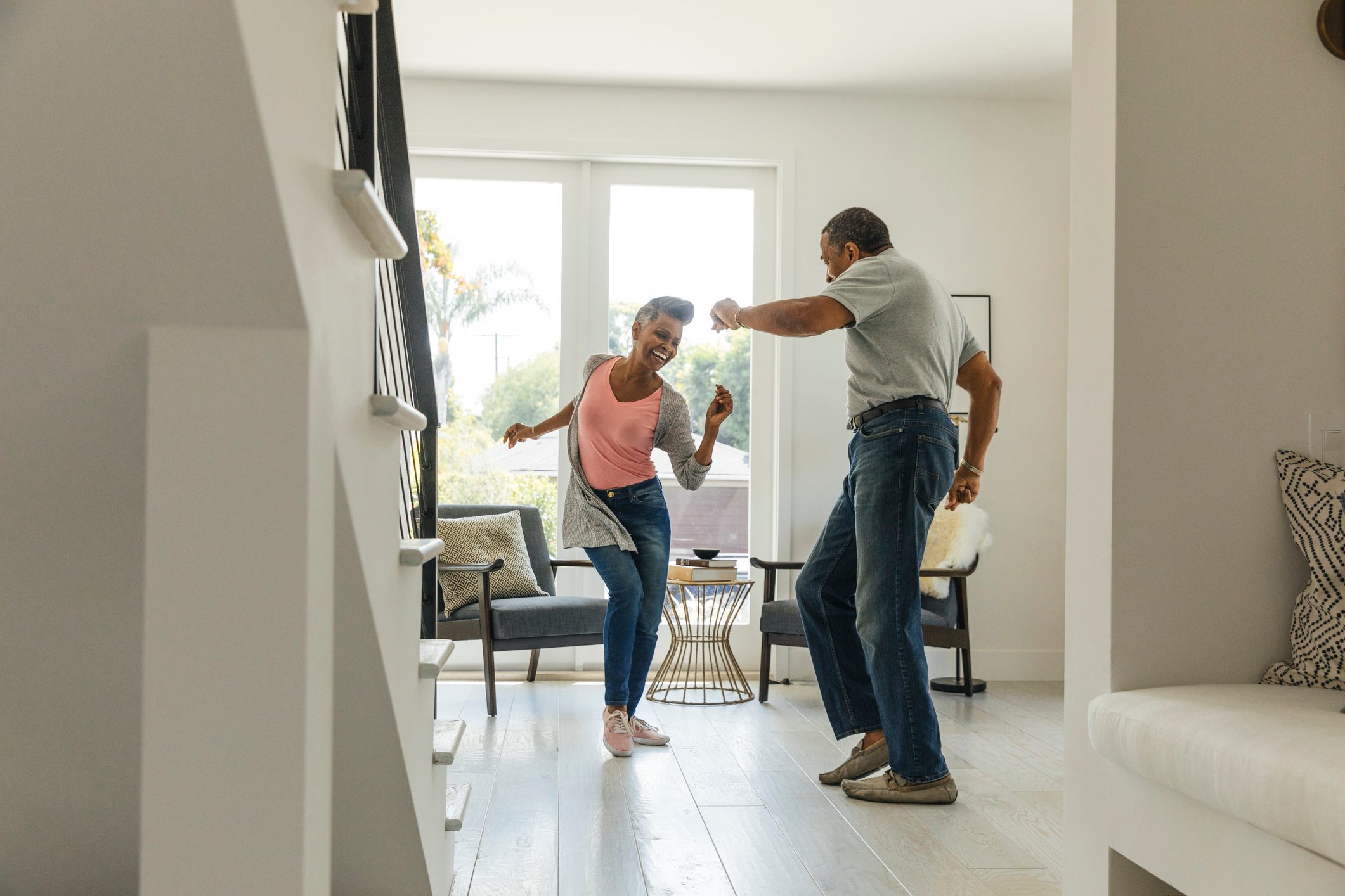 A couple dances joyfully in a bright, modern living room with large windows, chairs, and a small table, surrounded by greenery outside.