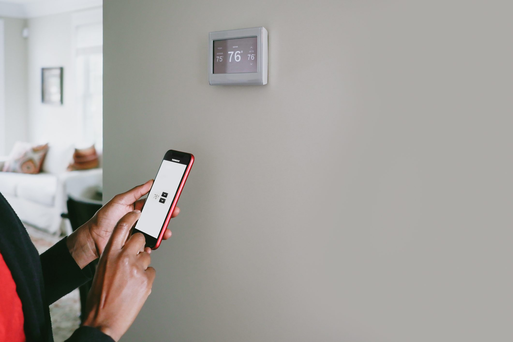 A person adjusts a thermostat using a smartphone, while a neutral-colored wall and a living room are visible in the background.