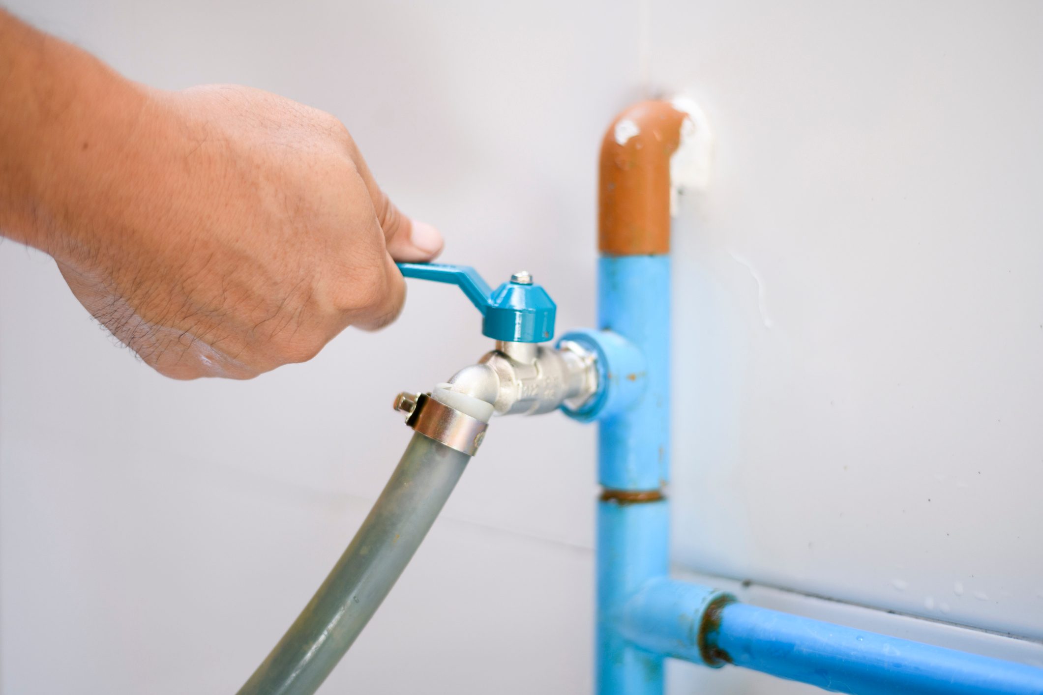 A hand turns a blue valve on a metal pipe, connecting to a green hose, in a minimalist environment with light-colored walls.