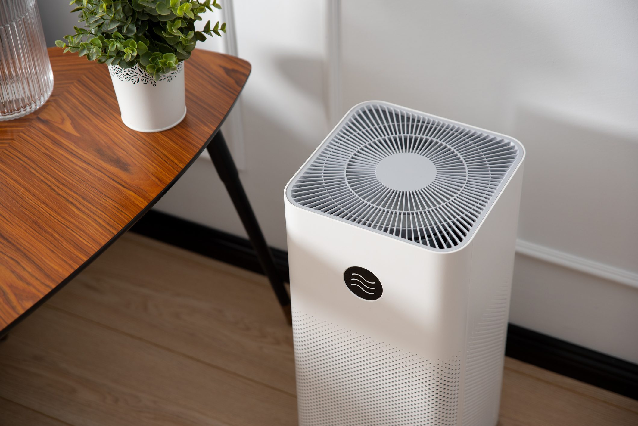 An air purifier stands on a wooden table, processing air, surrounded by a small plant and a glass container on a light wooden floor.