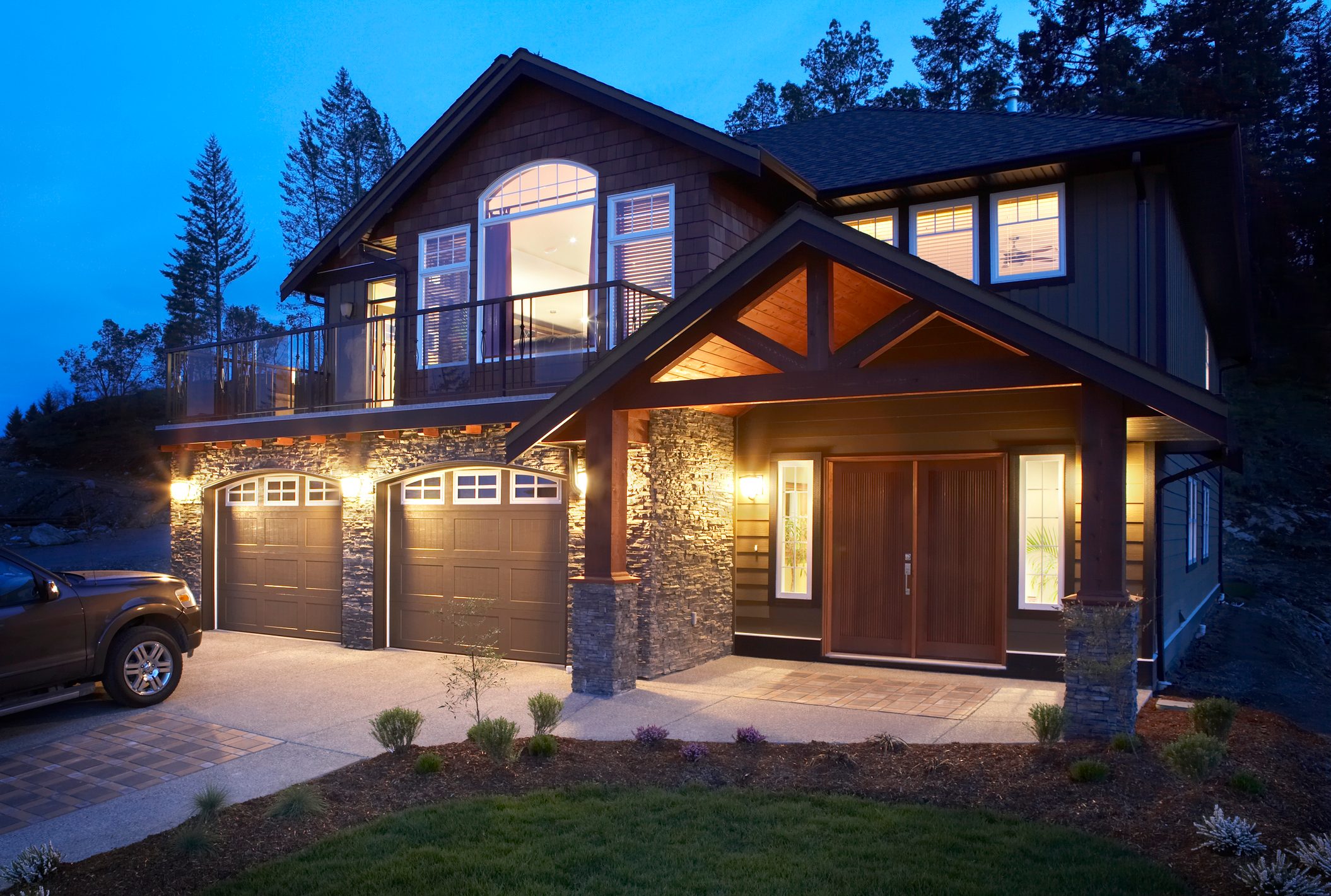 A modern house stands illuminated at dusk, featuring a stone façade, large windows, and a two-car garage, surrounded by a manicured lawn and trees.