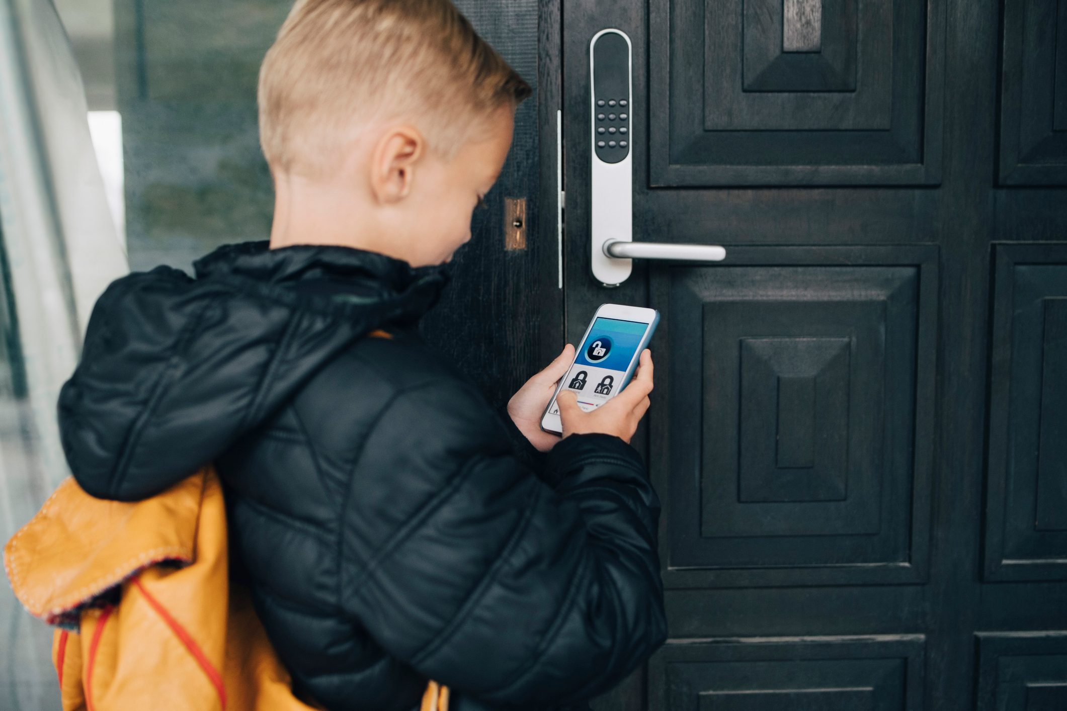 A boy holds a smartphone, attempting to unlock a modern door lock, while standing in front of a dark wooden door.
