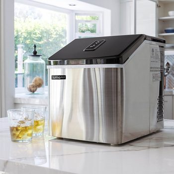 A stainless steel ice maker sits on a countertop, producing ice while two glasses filled with ice and drinks stand nearby in a bright kitchen.