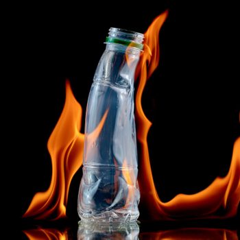 A plastic bottle stands upright while flames flicker around it, set against a dark background, creating a striking contrast.
