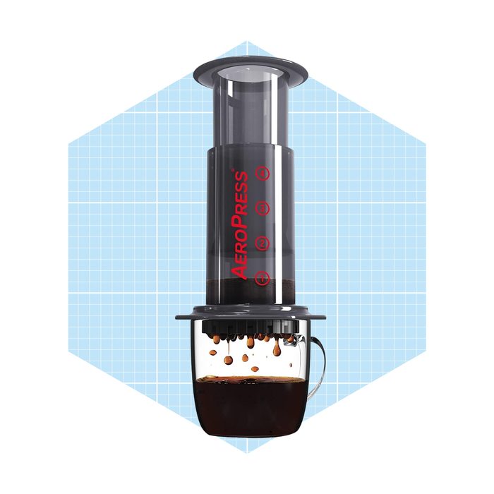 An Aeropress coffee maker extracts strong coffee, with brewed liquid dripping into a transparent cup below, all set against a light blue grid background.