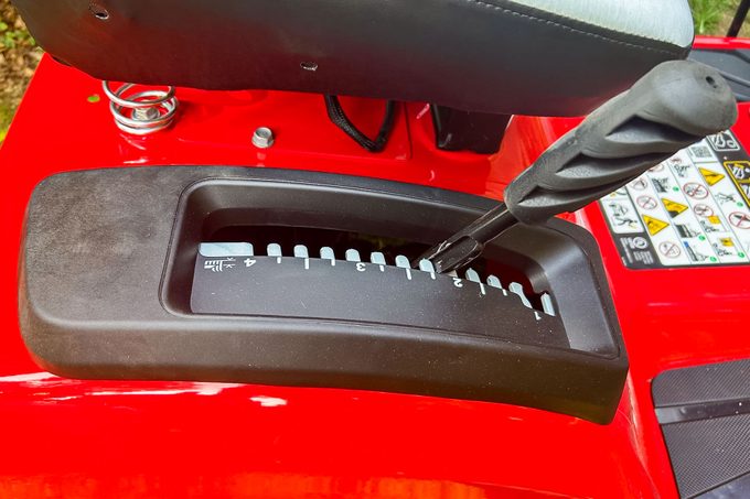 A black gear shift lever is positioned over a numbered console, indicating speed adjustments on a bright red lawn mower in a grassy context.