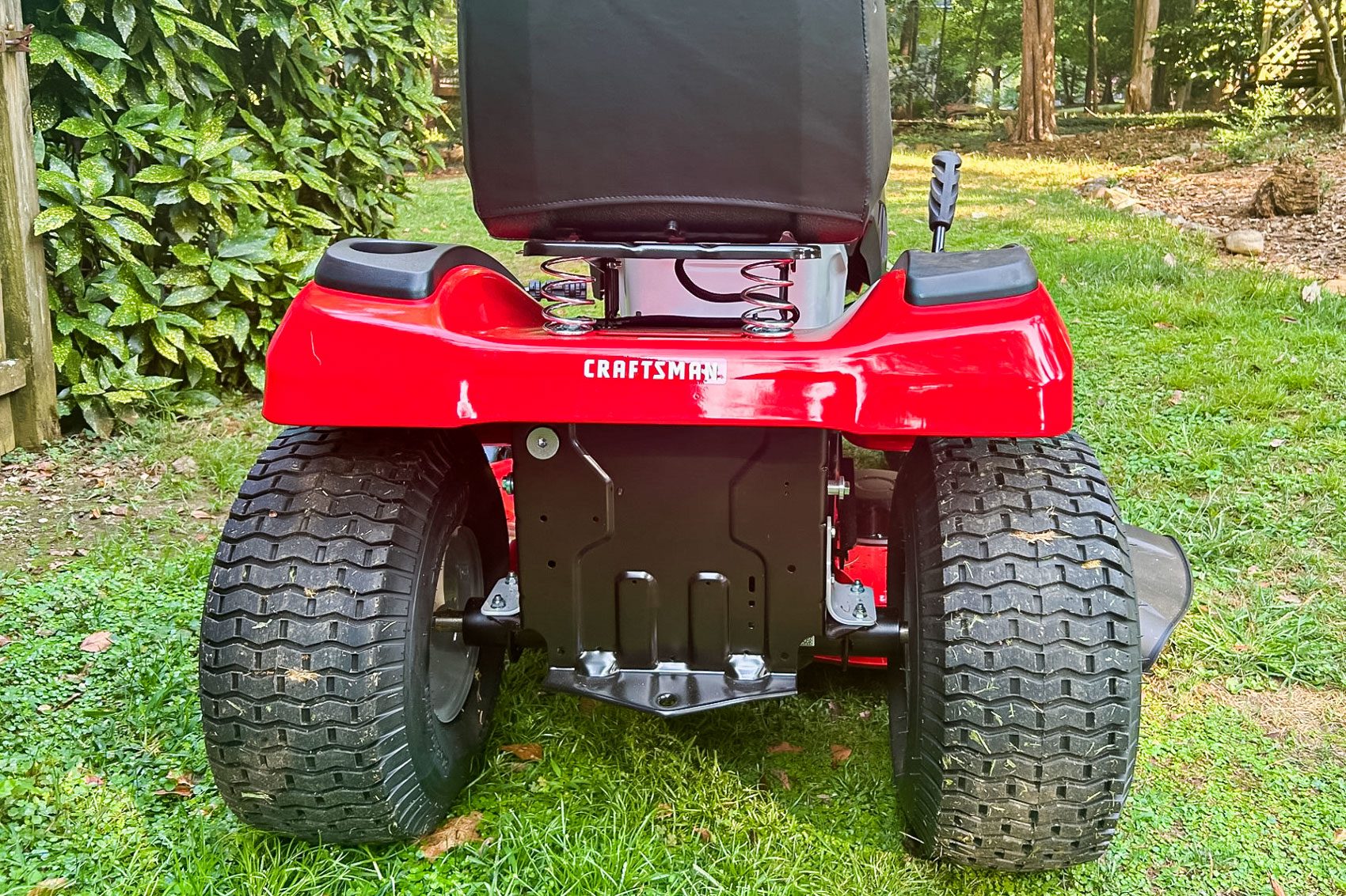A red lawn mower stands on green grass, with the rear view showing large wheels, a seat, and garden foliage in the background.