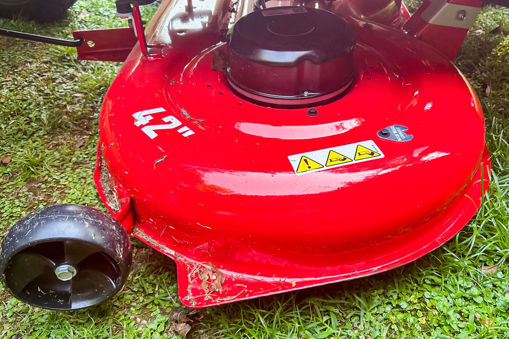 A red mower deck rests on green grass, displaying a 42-inch label, with a black wheel and warning symbols for safety.