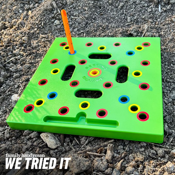 A green planting tool with holes and an orange measuring stick rests on dark soil, designed for precise seed spacing in gardening.