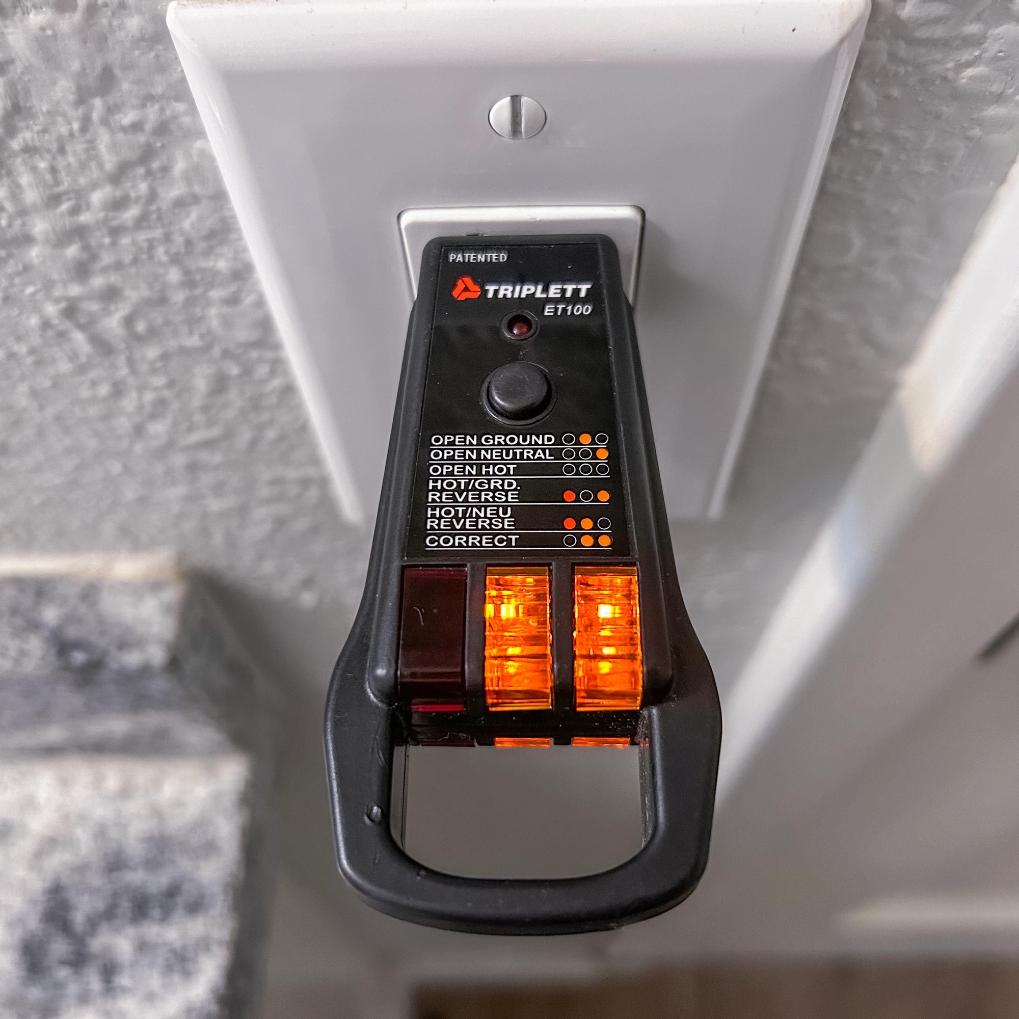 A voltage tester is plugged into a wall outlet, displaying lit indicators for electrical status in a room with a light-colored wall and steps visible.