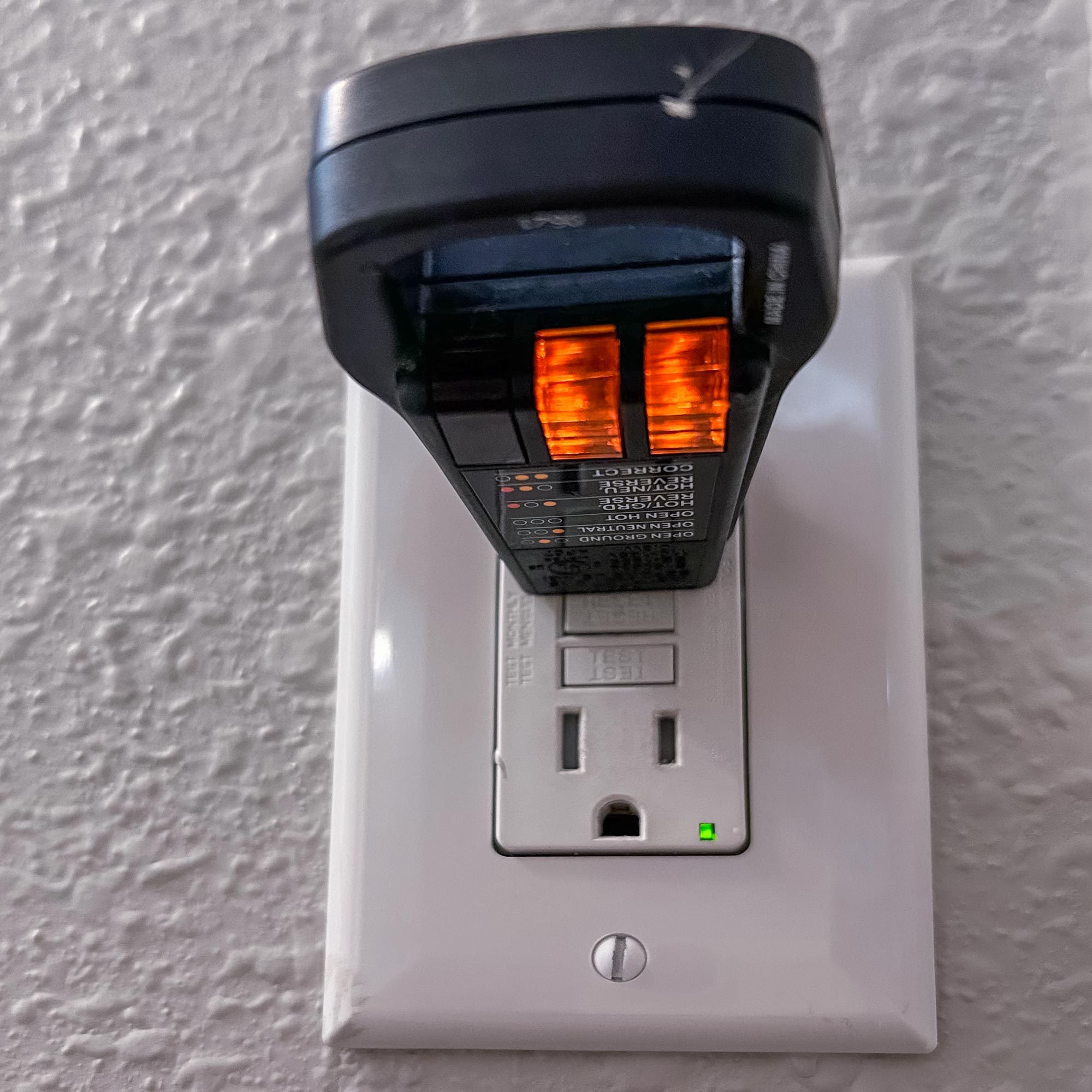 A voltage tester is plugged into a wall outlet, displaying orange lights, indicating testing. The background features a textured white wall.