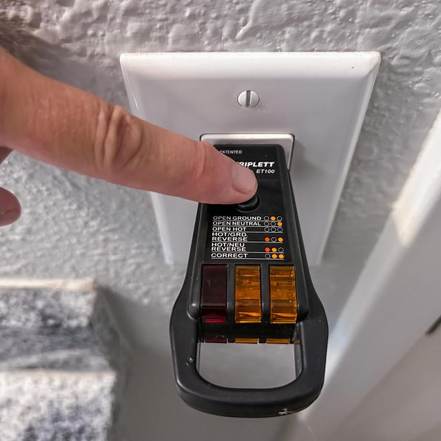 A hand presses a button on an electrical tester plugged into a wall outlet, indicating various electrical conditions with labeled lights.
