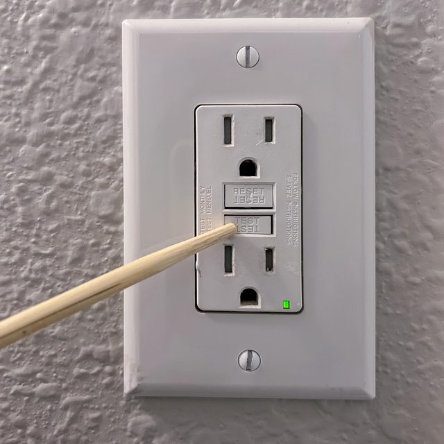 A test tool is applied to the "TEST" button on a white electrical outlet, situated on a textured gray wall, showing a green indicator light.