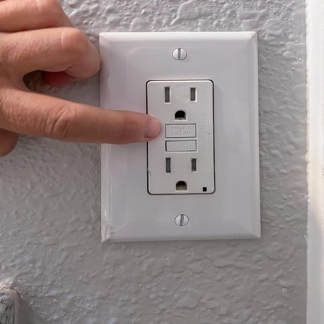 A hand presses the "RESET" button on a GFCI outlet mounted on a textured wall, indicating a potential electrical reset procedure.