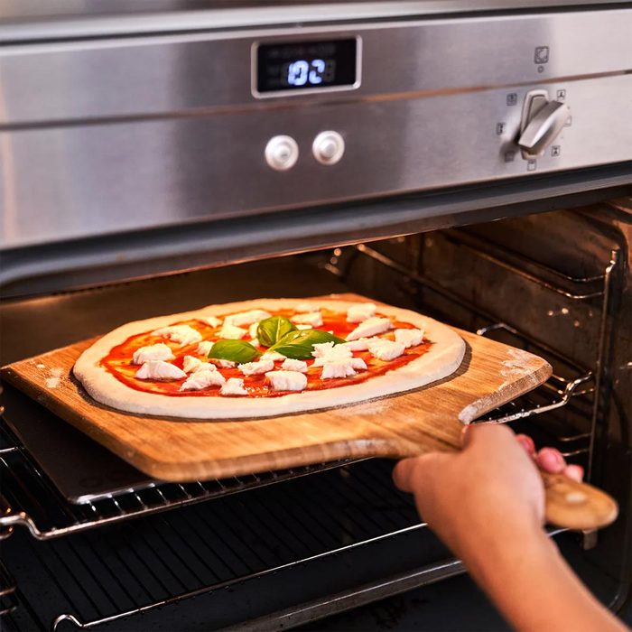 A hand pulls a wooden pizza peel from an oven, positioning a pizza topped with cheese and basil for baking in a modern kitchen.