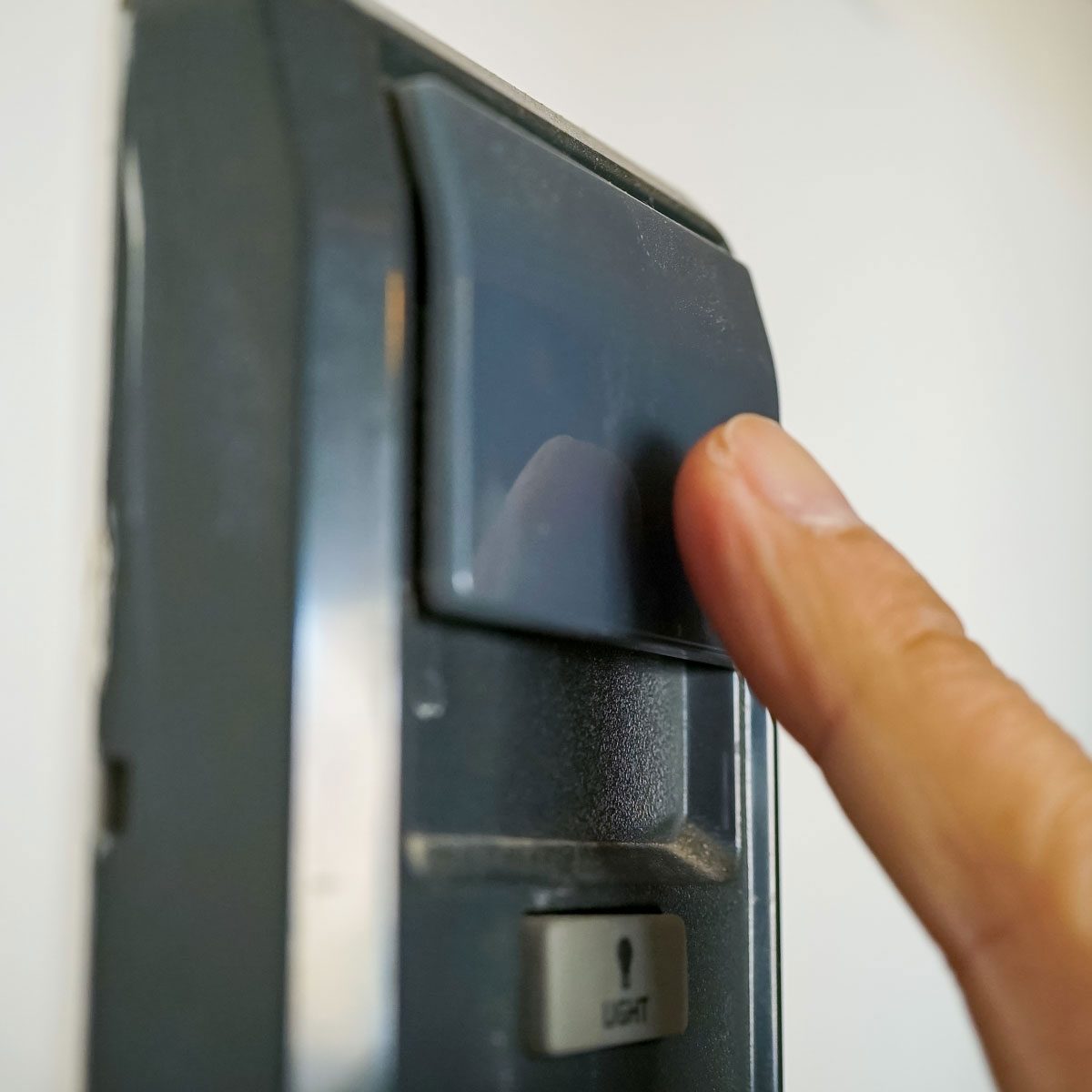 A finger presses a wall-mounted device, likely a switch, against a neutral-colored wall, indicating an action to activate lighting or another function.