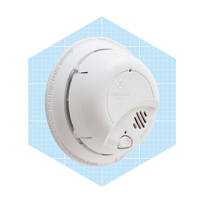 A round white smoke detector is mounted on a light blue, grid-patterned background, featuring a test button and air vents for smoke detection.