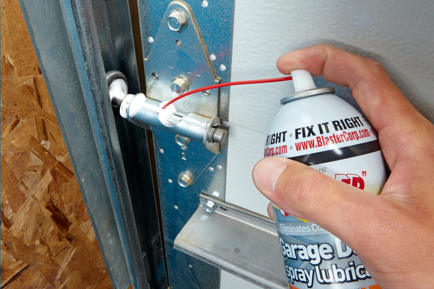 A hand holds a spray can, applying lubricant to a garage door hinge against a background of wood and metallic surfaces.