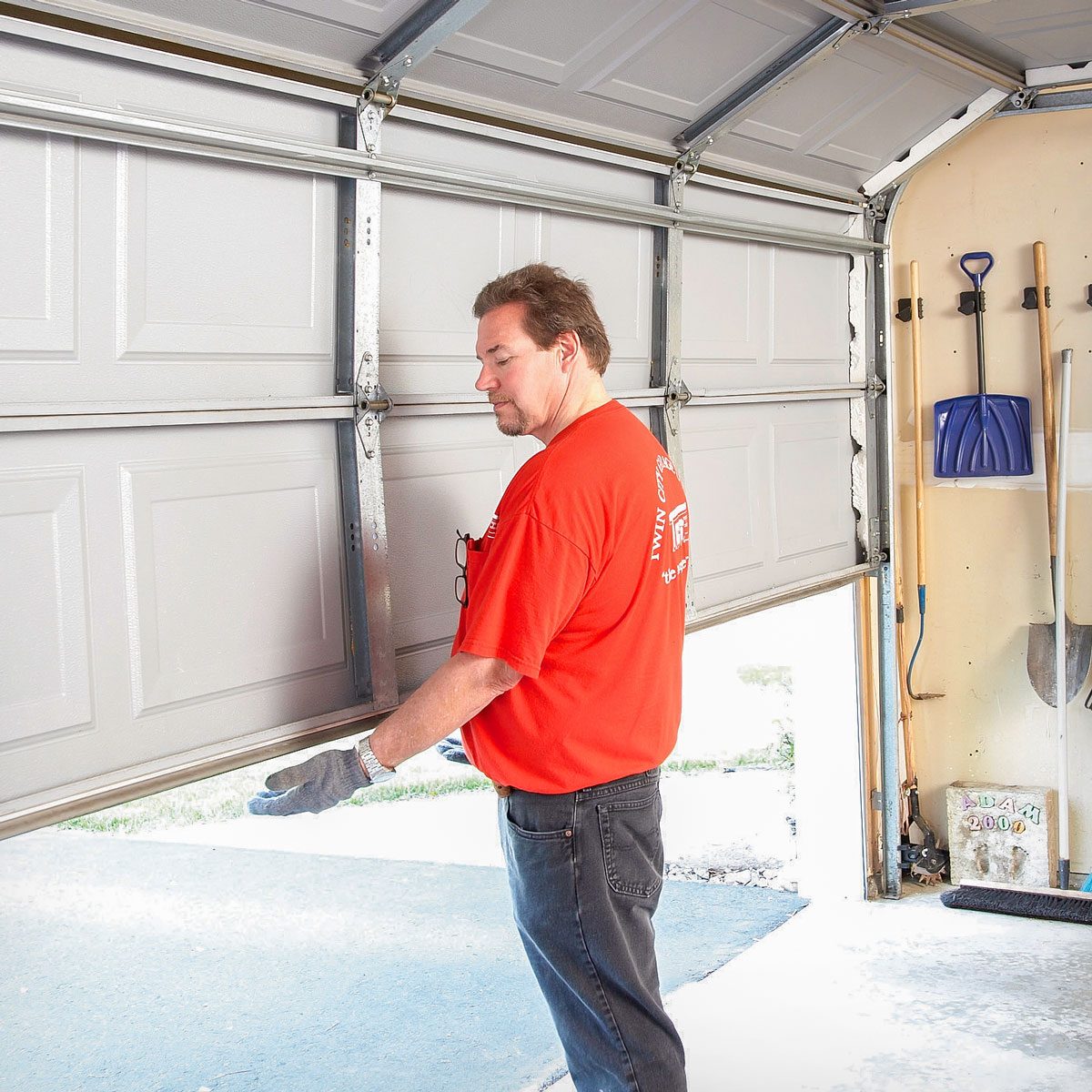 A man in a red shirt lifts a garage door, revealing an outdoor area. Tools are hung on a wall inside the garage.