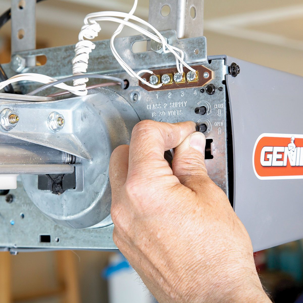 A hand adjusts settings on a garage door opener, surrounded by wires and mounted on a metal frame in a well-lit garage environment.