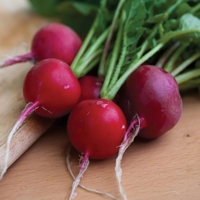 Red radishes with green leaves rest on a wooden surface, showcasing their glossy skin and delicate roots. The arrangement is fresh and appetizing.